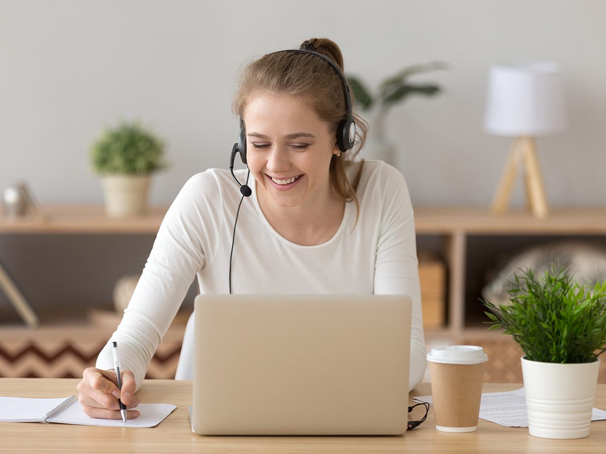 Smiling woman wearing headset writing notes studying online on laptop