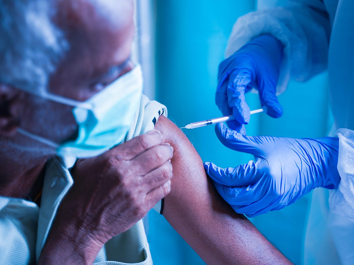 Senior elderly patient with mask getting vaccination or jab at hospital - Docotor in protective Hazmat suit treating Covid patient injecting anti-viral vials at covid care Centre.
