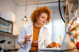 Smiling young biochemist using laptop at her desk in laboratory. Young focused red haired chemist working on scientific research while using computer in a laboratory.